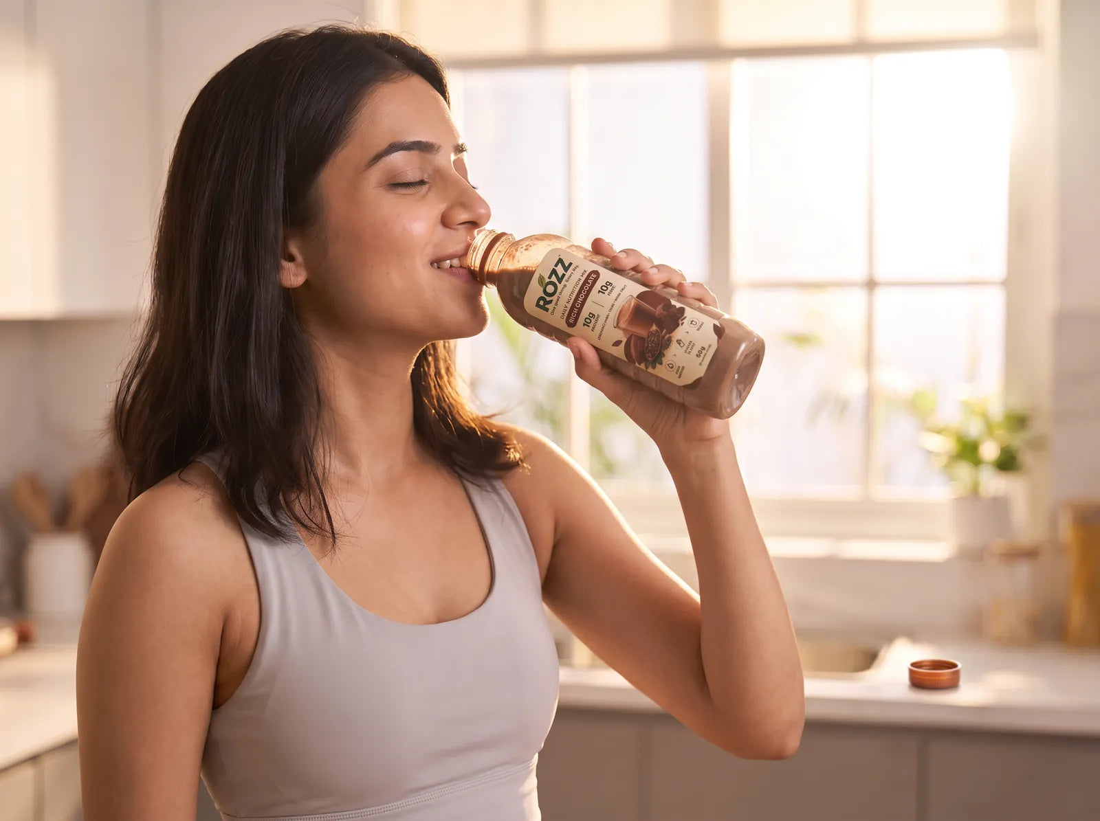 A woman drinking ROZZ Chocolate from the bottle in a sunlit kitchen